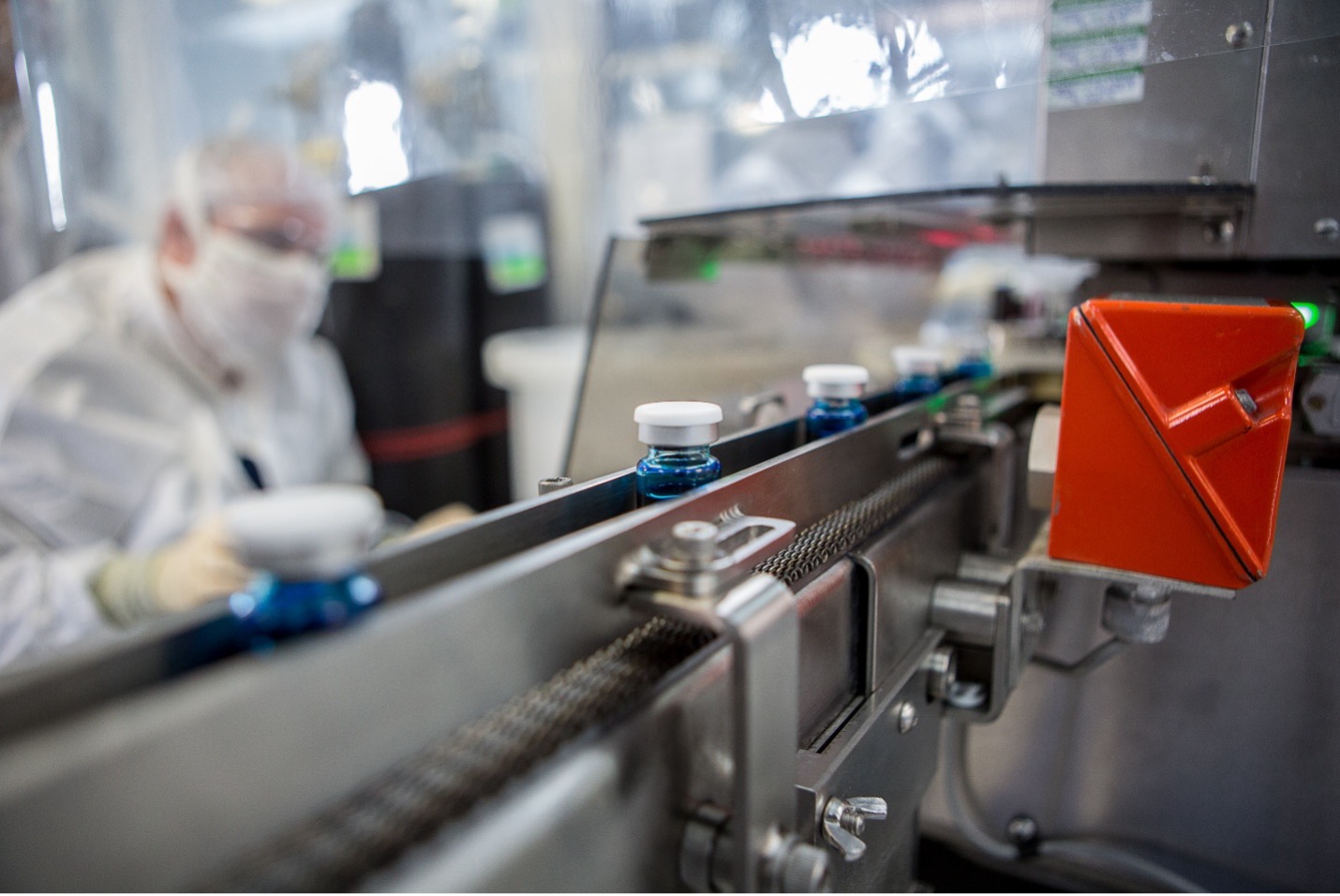 A close-up view of an automated production line in a sterile facility, showing small glass vials filled with blue liquid moving along a metal conveyor, while a worker in protective clothing and gloves is blurred in the background.