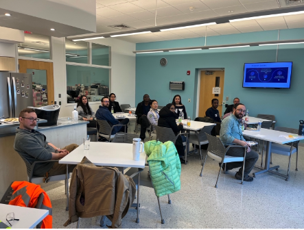 Group of employees seated at tables in a bright breakroom-style meeting space, looking toward the camera.