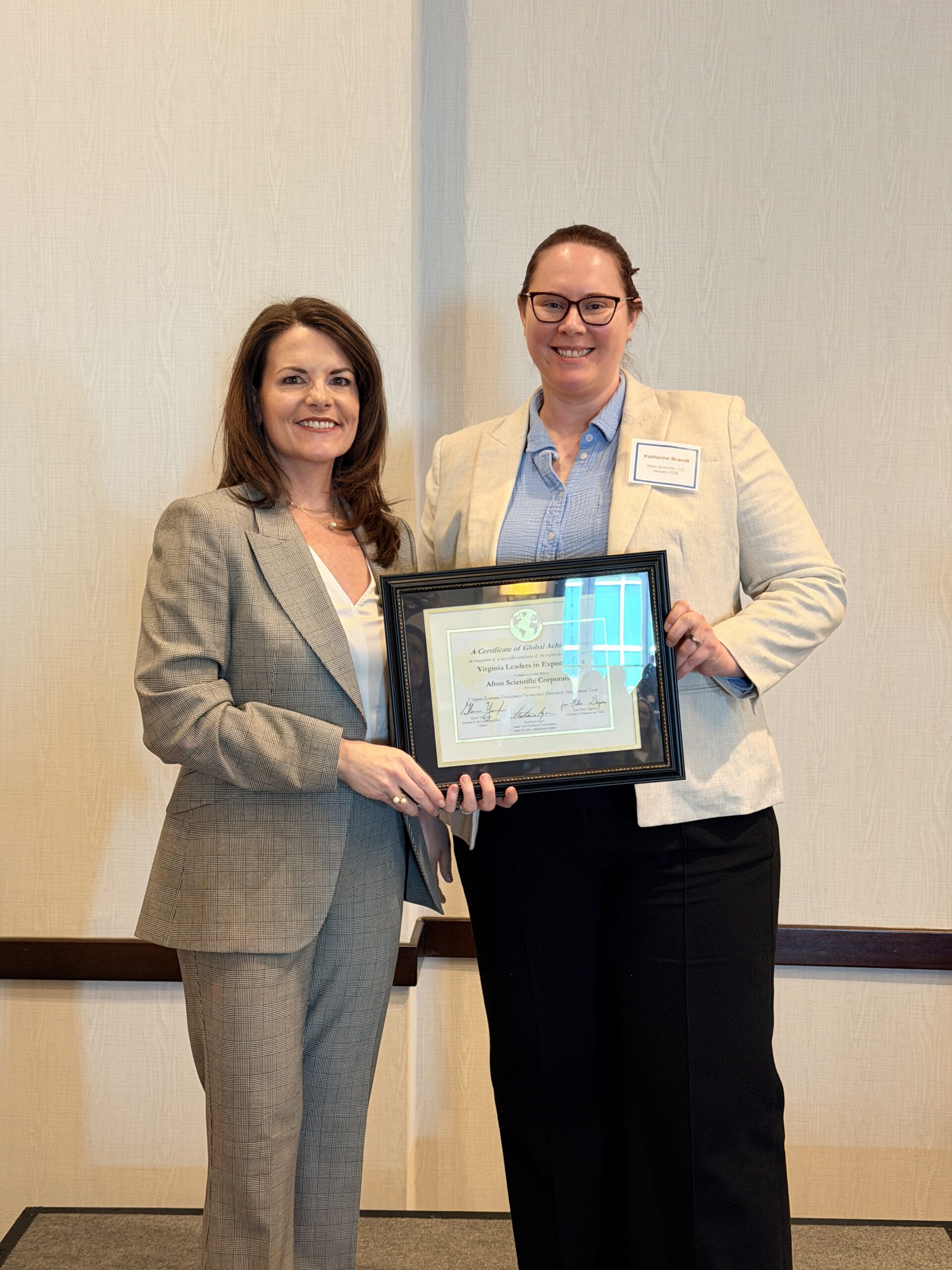 Two women in business attire stand side by side indoors, holding a framed certificate together and facing the camera. Their faces are blurred, and they are positioned against a plain light-colored wall, suggesting a formal award or recognition event.